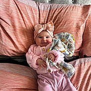 Brianna is registered to the contest to win money with this photo: baby, infant, pink_clothing, headband, bow, blanket, bed, pillow, striped, knitwear, smile, cute, child, person, indoors, soft_texture, cozy, portrait, young, happy