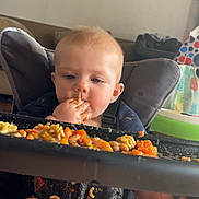 Zachery is registered to the contest to win money with this photo: baby, messy, food, pasta, high_chair, eating, child, indoor, meal, hands, face, legs, tray, living_room, furniture, clothing, expression, closeup, seat, background