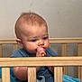baby, child, crib, wooden_crib, blue_shirt, bear_print, blanket, indoor, sitting, thumb_sucking, infant, cute, face, head, person, young_child, toy, room, wall, expression