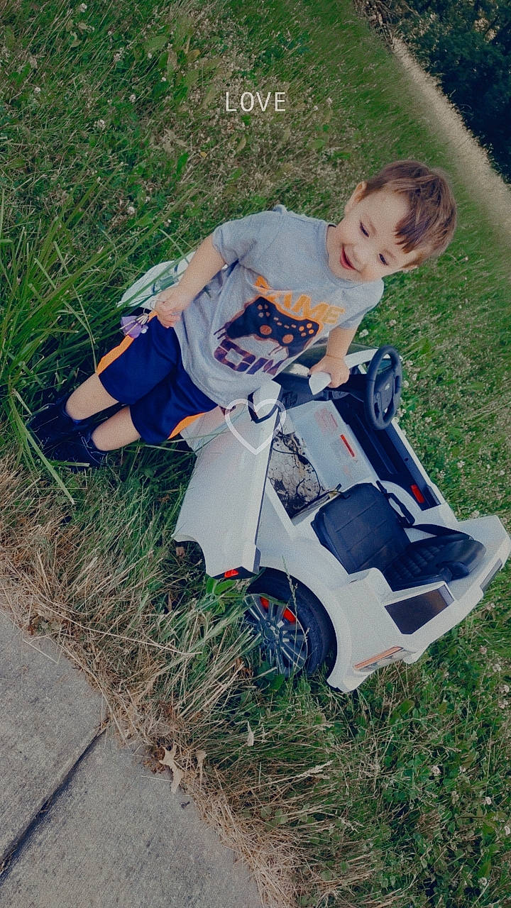 Ashton is registered to the contest to win money with this photo: automotive_wheel_system, baby_products, baby_toddler_clothing, electric_blue, face, grass, happy, head, joy, leisure, people_in_nature, person, plant, recreation, riding_toy, sitting, smile, tire, toddler, tree