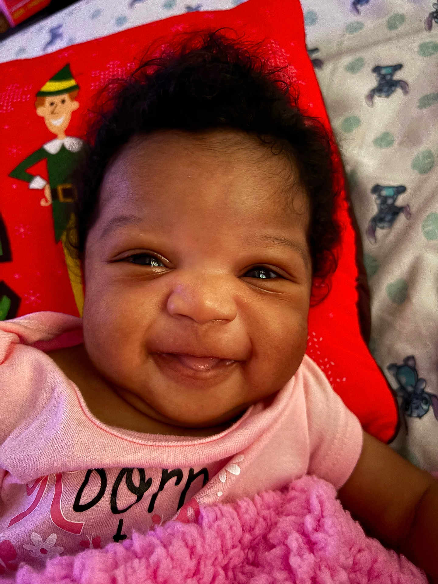 Kharia joined the competition — help win amazing prizes! baby, smile, face, pink_clothing, blanket, pillow, curly_hair, infant, happy, cute, child, portrait, indoor, cozy, red_pillow, illustration, closeup, skin, person, expression