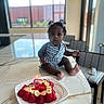 Tianna joined the competition — help win amazing prizes! baby, birthday_cake, candle, child, curly_hair, fruit, indoor, kitchen, marble_countertop, plate, person, sitting, strawberry, table, toddler, window, young_child, striped_clothing, celebration, food