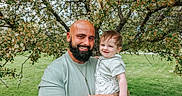 Waylan joined the competition — help win amazing prizes! baby, beard, casual_clothing, child, daylight, family, flower, grass, green, happy, holding, man, nature, outdoor, parent, person, portrait, smile, tattoo, tree