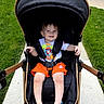 Waylan joined the competition — help win amazing prizes! toddler, child, stroller, outdoor, sidewalk, grass, happy, smile, orange_shorts, tshirt, footwear, baby, seatbelt, sunny_day, person, walking_path, park, casual_clothing, baby_stroller, summer