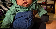 Jruziah is registered to the contest to win money with this photo: adult_hand, baby, blanket, blue_jacket, child, clothing, curious_expression, cute, face, furniture, gloves, green_jacket, hooded_jacket, indoor, living_room, person, portrait, sitting, television, warm_clothing