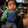 adult_hand, baby, blanket, blue_jacket, child, clothing, curious_expression, cute, face, furniture, gloves, green_jacket, hooded_jacket, indoor, living_room, person, portrait, sitting, television, warm_clothing