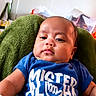 baby, blue_shirt, child, clothing, cushion, cute, expression, face, green, head, indoor, infant, person, portrait, relaxed, seated, skin, text_on_clothing, toddler, young_child