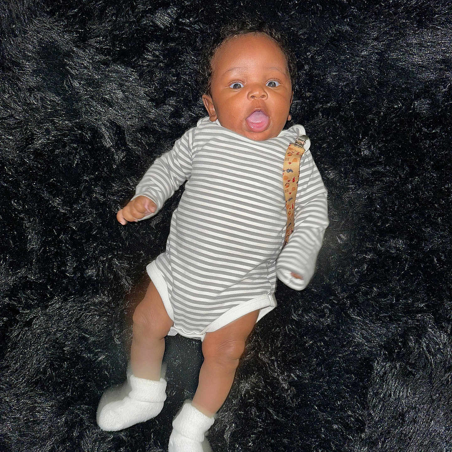 Terrance Iii is registered to the contest to win money with this photo: adorable, baby, black_fur_rug, child, clothing, cute, face, head, human, indoors, infant, limbs, lying_down, newborn, portrait, small_person, soft_texture, striped_onesie, surprised_expression, white_socks