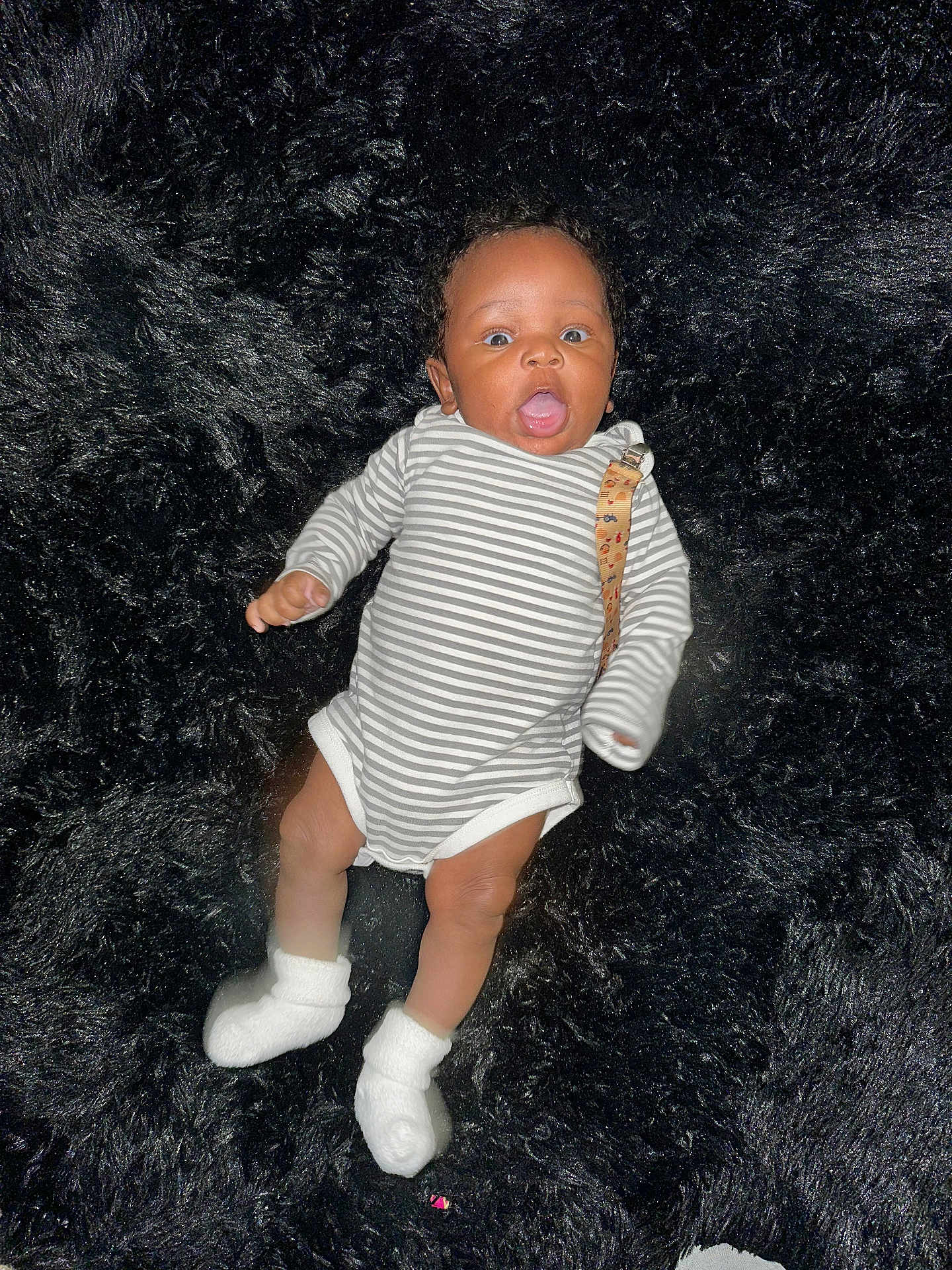 Terrance Iii is registered to the contest to win money with this photo: baby, infant, striped_onesie, white_socks, black_fur_rug, lying_down, surprised_expression, child, cute, soft_texture, indoors, small_person, human, adorable, newborn, face, head, limbs, clothing, portrait