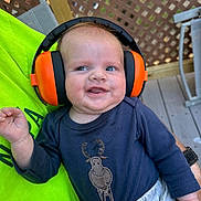 Joey is registered to the contest to win money with this photo: baby, child, ear_protectors, orange, blue_shirt, moose_graphic, gray_pants, smiling, outdoor, deck, wooden_lattice, chair, person, infant, happy, cute, casual_clothing, sitting, daylight, closeup