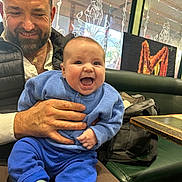 Hugo a rejoint le concours — aidez-le/la à gagner de superbes lots ! baby, man, beard, blue_clothing, sweater, pants, smile, indoor, restaurant, booth, table, hand, glass_window, poster, artwork, bag, casual, seated, joyful, person