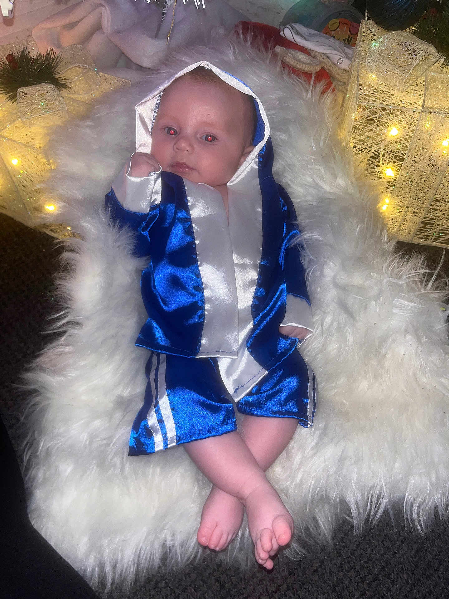 Tommie joined the competition — help win amazing prizes! baby, infant, blue_robe, boxing_robe, fluffy_rug, crossed_legs, feet, face, red_eye, indoor, carpet, decorations, lights, holiday, soft_texture, cute, child, small_hands, portrait, relaxed
