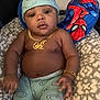 baby, child, smiling_face, hat, pants, gold_necklace, gold_bracelet, spiderman_pillow, cushion, patterned_fabric, indoor, person, portrait, baby_clothing, accessory, relaxing, furniture, cute, young_child, seated