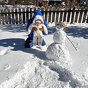Alexandra joined the competition — help win amazing prizes! baby, person, snowman, snow, winter, fence, blue_hoodie, hat, snowy_backyard, stick_arms, baseball_cap, cold_weather, outdoor, child, smile, white_suit, sunlight, footprints, house, tree