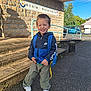 child, boy, smile, backpack, preschool, school_sign, jacket, sneakers, steps, stone_wall, outdoor, pavement, railing, sunlight, portrait, clothing, car, trees, happy, sidewalk