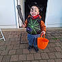 child, costume, halloween, face_paint, spider_headband, foam_club, orange_bucket, doormat, doorway, pavement, jacket, smile, shoes, trick_or_treat, outdoor, porch, toddler, bucket, happy, decor