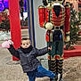 child, nutcracker, toy, pink_plush, holiday_display, christmas_decoration, festive_lights, outdoor, portrait, surprised_expression, jacket, scarf, sneakers, gravel, statue, tree, decorations, night_scene, person, playful