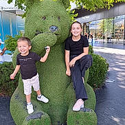 Tim a rejoint le concours — aidez-le/la à gagner de superbes lots ! child, boy, girl, topiary_bear, green_sculpture, statue, shopping_mall, tree, outdoor, smile, happiness, casual_clothing, sneakers, pavement, bushes, public_art, tourists, daylight, photo_op, playful_pose