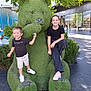 child, boy, girl, topiary_bear, green_sculpture, statue, shopping_mall, tree, outdoor, smile, happiness, casual_clothing, sneakers, pavement, bushes, public_art, tourists, daylight, photo_op, playful_pose
