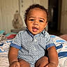 baby, child, infant, curly_hair, blue_shirt, patterned_clothing, cushion, whale_pattern, smiling, indoors, portrait, cute, sitting, hands, face, eyes, mouth, skin, hair, background_blur