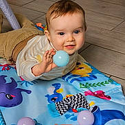 Edzio participe au concours pour gagner de l'argent avec cette photo : baby, child, play_mat, toy_ball, floor_tiles, indoor, happy, smiling, clothing, striped_shirt, pants, hand, face, head, blue_eyes, cute, teething_toy, colorful, playtime, lying_down