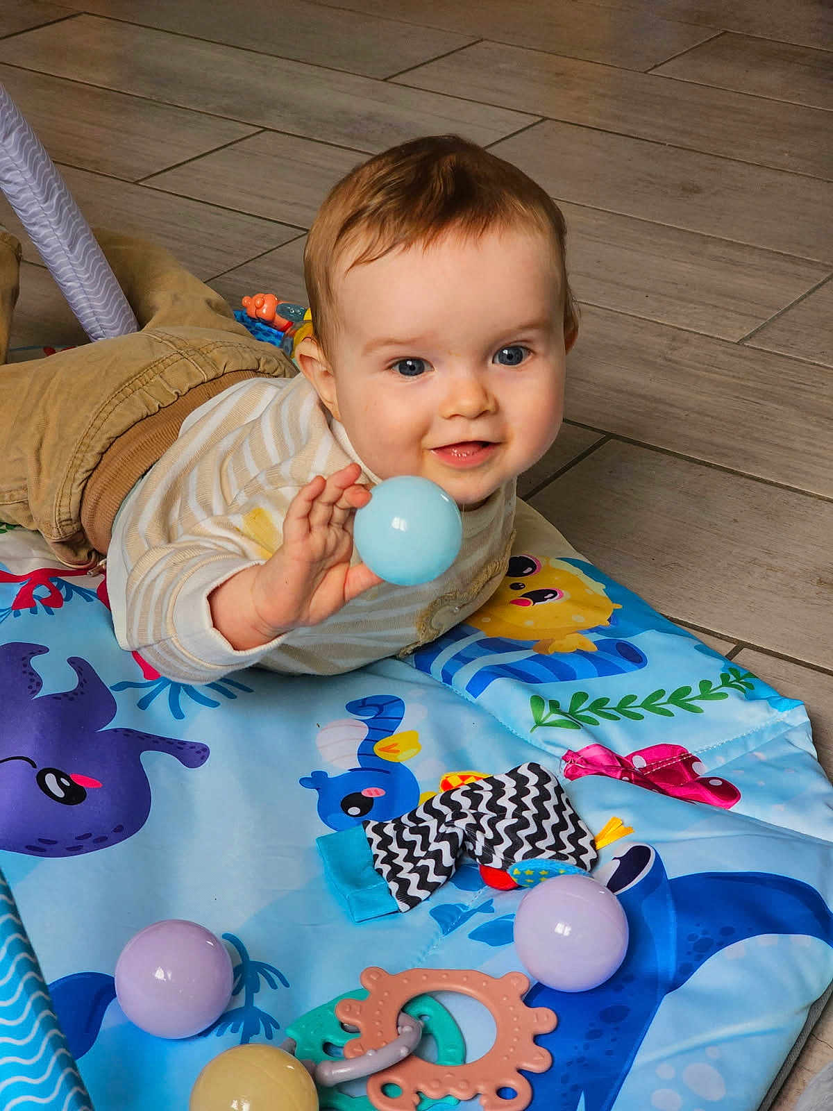 Edzio participe au concours pour gagner de l'argent avec cette photo : baby, child, play_mat, toy_ball, floor_tiles, indoor, happy, smiling, clothing, striped_shirt, pants, hand, face, head, blue_eyes, cute, teething_toy, colorful, playtime, lying_down