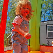 Nova is registered to the contest to win money with this photo: child, toddler, denim_overalls, boots, bounce_house, inflatable, outdoor, sunny, daylight, trees, blue_sky, grass, play, fun, smile, hair, portrait, person, happy, recreation