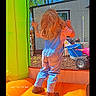 child, blonde_hair, denim_overalls, boots, bounce_house, inflatable, yellow, orange, outdoor, backyard, toy_vehicle, mesh_wall, sunny, daytime, grass, shed, play, standing, casual_clothing, timestamp