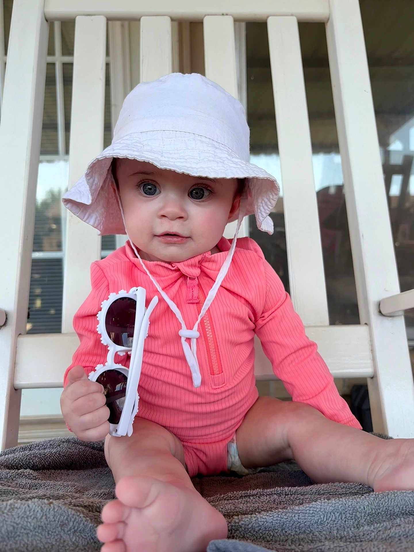 Hazel is registered to the contest to win money with this photo: baby, child, pink_outfit, white_hat, sunglasses, chair, towel, foot, hand, face, indoor, curious, sitting, portrait, cute, infant, clothing, headwear, person, closeup