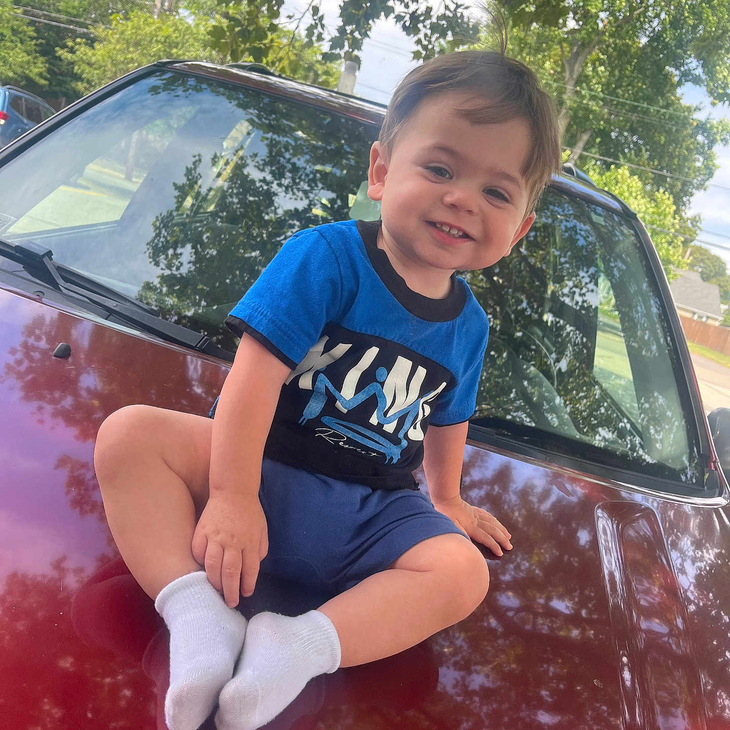 Myles Greco joined the competition — help win amazing prizes! blue_shirt, car, casual_clothing, child, daylight, grass, happy, hood, nature, outdoor, parked_car, portrait, reflection, sidewalk, smile, sunlight, toddler, trees, white_socks, young_child