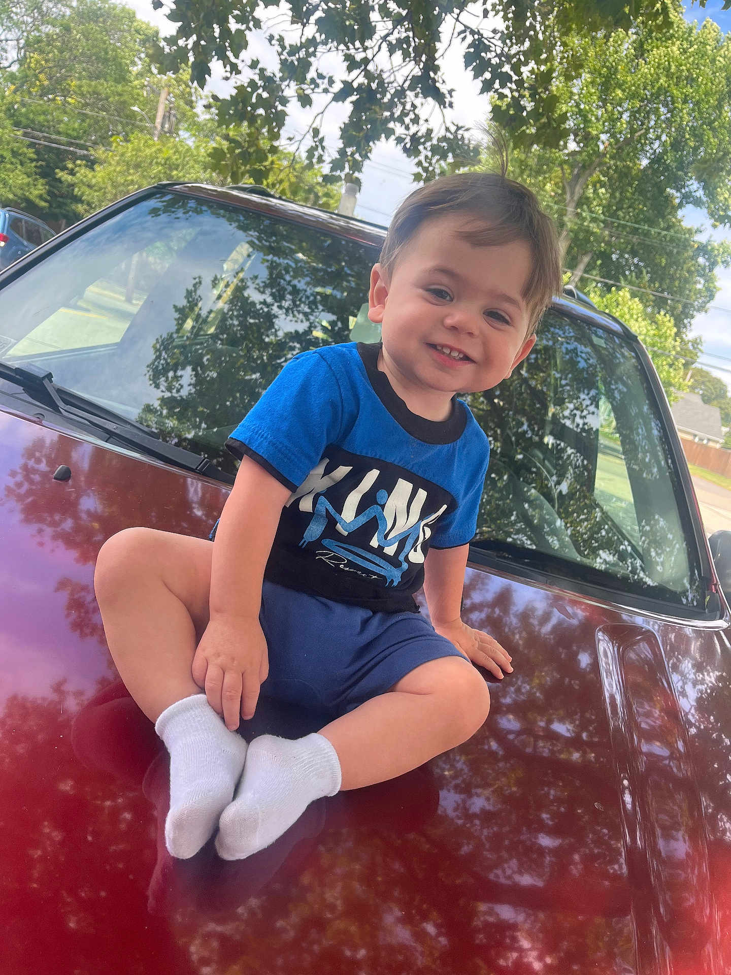 Myles Greco joined the competition — help win amazing prizes! toddler, child, smile, blue_shirt, white_socks, car, hood, outdoor, daylight, trees, reflection, grass, happy, casual_clothing, portrait, young_child, sidewalk, nature, parked_car, sunlight