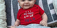 Easton is registered to the contest to win money with this photo: baby, infant, smile, red_onesie, socks, cushion, blanket, indoors, cute, child, happy, portrait, sitting, pattern, fabric, face, skin, small_person, comfort, home