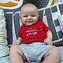 baby, infant, smile, red_onesie, socks, cushion, blanket, indoors, cute, child, happy, portrait, sitting, pattern, fabric, face, skin, small_person, comfort, home