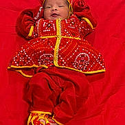 Reyna joined the competition — help win amazing prizes! baby, infant, red_clothing, traditional_outfit, lying_down, colorful, cute, child, face, headwear, footwear, patterned_clothing, fabric, soft, small_hands, portrait, indoors, celebration, bright_colors, warm