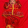 baby, infant, red_clothing, traditional_outfit, lying_down, colorful, cute, child, face, headwear, footwear, patterned_clothing, fabric, soft, small_hands, portrait, indoors, celebration, bright_colors, warm