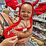 baby, child, devil_hat, red, smiling, person, holding, store, indoor, kitchenware, home_goods, shelves, adult, patterned_dress, happy, cute, costume, retail, shopping, headwear