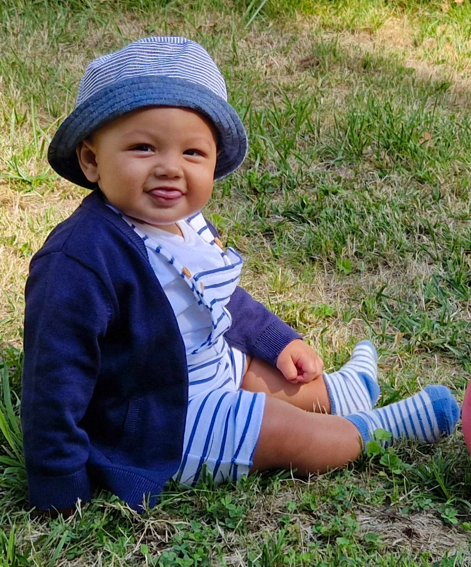 Colin participe au concours pour gagner de l'argent avec cette photo : baby, child, grass, hat, striped_clothing, cardigan, socks, tongue_out, outdoor, cute, smiling, sitting, nature, sunlight, happy, person, infant, casual_wear, summer, playful