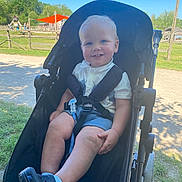 Jayden joined the competition — help win amazing prizes! toddler, child, stroller, outdoor, park, sunny, grass, playground, smile, blue_eyes, shorts, shoes, white_shirt, daylight, happy, sitting, nature, fence, trees, shade