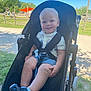 toddler, child, stroller, outdoor, park, sunny, grass, playground, smile, blue_eyes, shorts, shoes, white_shirt, daylight, happy, sitting, nature, fence, trees, shade