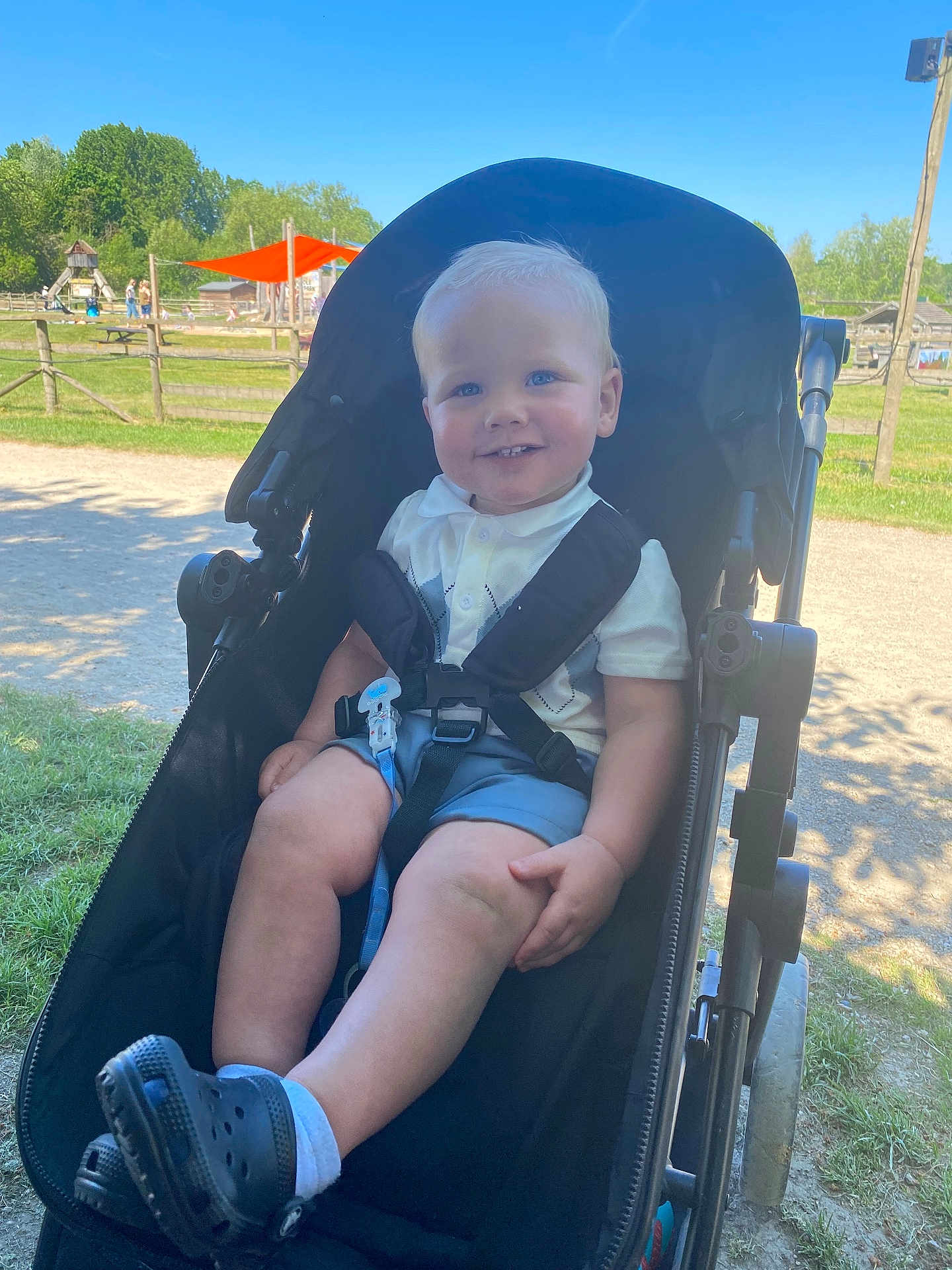 Jayden joined the competition — help win amazing prizes! toddler, child, stroller, outdoor, park, sunny, grass, playground, smile, blue_eyes, shorts, shoes, white_shirt, daylight, happy, sitting, nature, fence, trees, shade