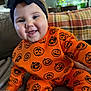 Brynlee is registered to the contest to win money with this photo: baby, child, smiling, orange_outfit, jack_o_lantern, headband, black_bow, couch, plaid_blanket, indoors, happy, sitting, cute, face, hand, cozy, clothing, person, portrait, home