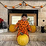 toddler, child, pumpkin_outfit, orange_clothing, black_headband, fireplace, pumpkins, halloween_decor, skeletons, black_cat_figurine, mirror, flowers, autumn_leaves, spell_book, carpet, indoor, smiling, festive, holiday, decorations