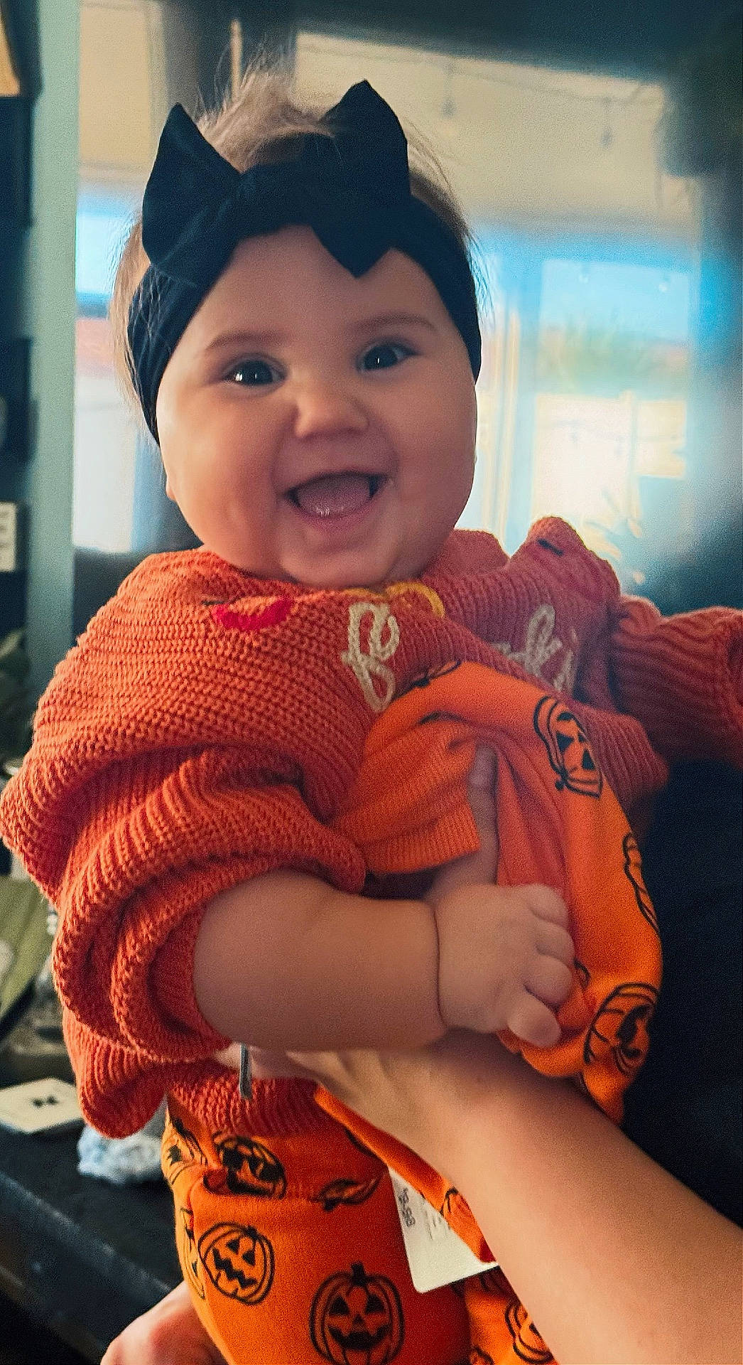 baby, black_headband, child, cozy, cute, face, festive, hand, happy, headband, holding, indoor, orange_clothing, person, portrait, pumpkin_pattern, smiling, sweater, window, young_child
