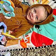 Hudson is registered to the contest to win money with this photo: baby, child, hoodie, brown_clothing, smile, face, toy, playmat, carpet, nasal_cannula, infant, lying_down, colorful, patterned, cute, indoor, happy, person, plush, wearing_hood
