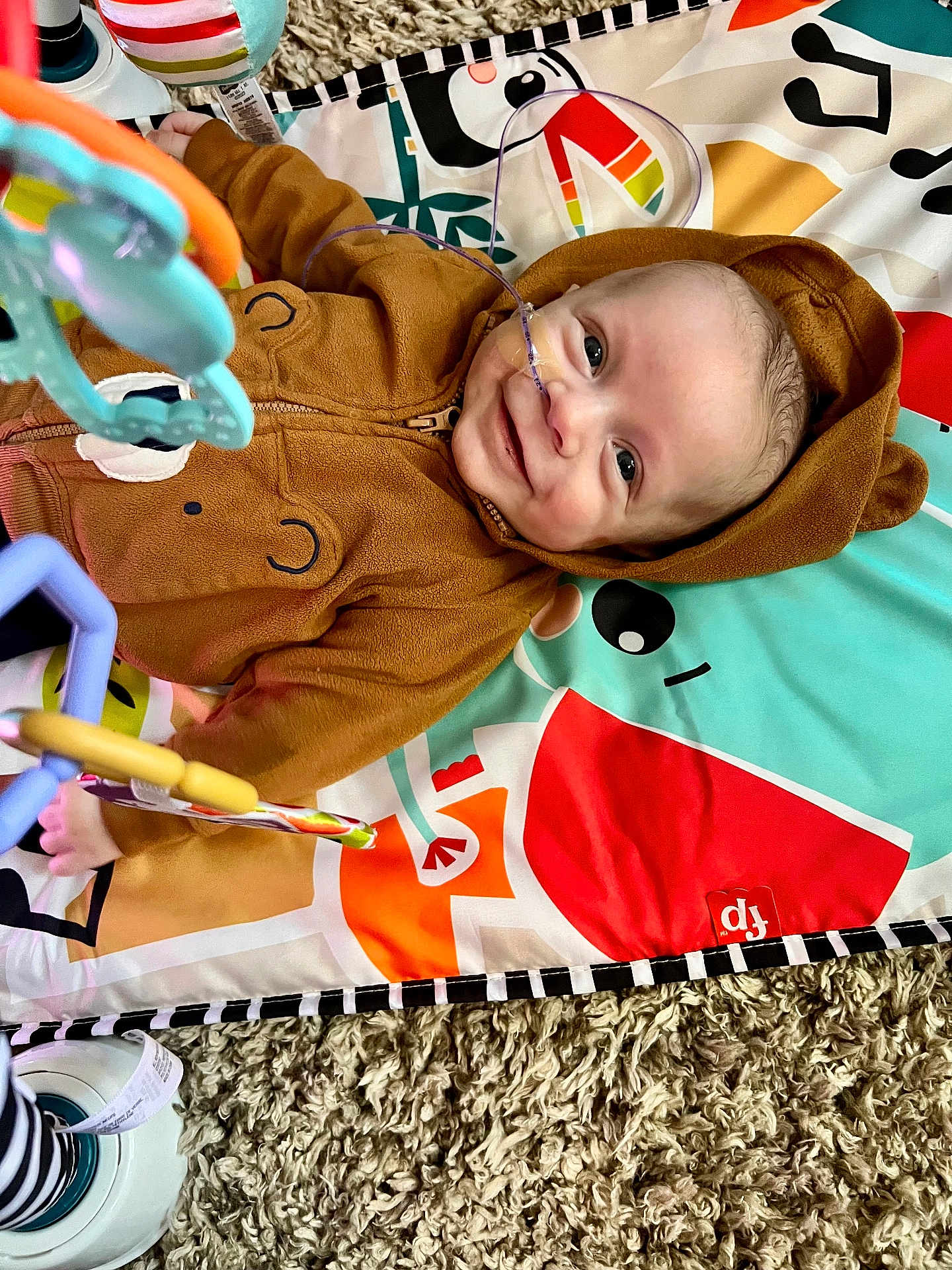 Hudson is registered to the contest to win money with this photo: baby, child, hoodie, brown_clothing, smile, face, toy, playmat, carpet, nasal_cannula, infant, lying_down, colorful, patterned, cute, indoor, happy, person, plush, wearing_hood