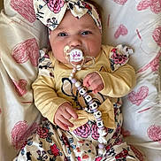 Maria a rejoint le concours — aidez-le/la à gagner de superbes lots ! baby, infant, pacifier, headband, bow, floral_clothing, blanket, heart_pattern, cute, child, hand, face, expression, indoors, person, clothing, lying_down, pink, yellow, comfort