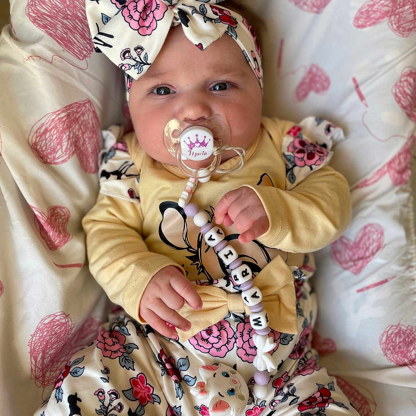 Maria a rejoint le concours — aidez-le/la à gagner de superbes lots ! baby, infant, pacifier, headband, bow, floral_clothing, blanket, heart_pattern, cute, child, hand, face, expression, indoors, person, clothing, lying_down, pink, yellow, comfort