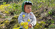 Lisandro participe au concours pour gagner de l'argent avec cette photo : toddler, child, forest, nature, moss, rocks, green_pants, hoodie, dinosaur_hood, yellow_flowers, outdoor, sitting, casual_clothing, curious_expression, daylight, young_child, playful, scenic, flora, leaves