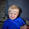child, boy, blond_hair, smiling, grinning, blue_shirt, portrait, indoors, living_room, couch, rug, furniture, seated, toy, holding_object, happy, face, head, arm, casual_clothing