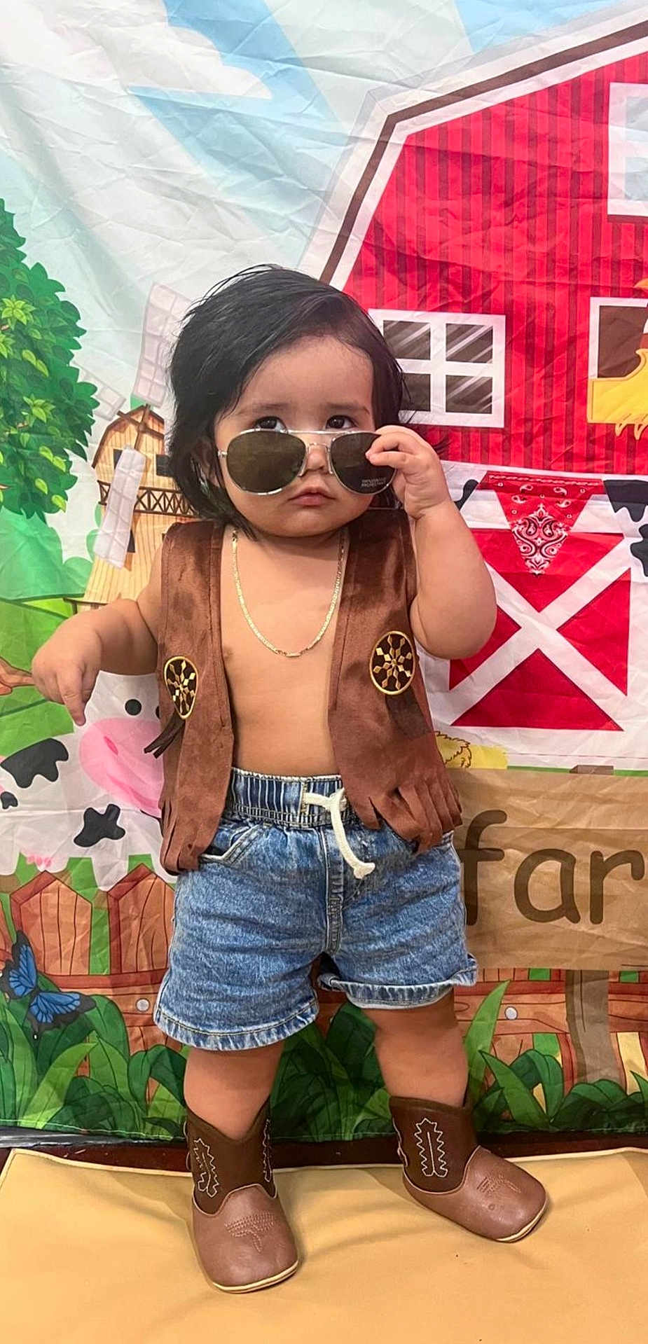 Ian is registered to the contest to win money with this photo: child, boy, sunglasses, vest, denim_shorts, cowboy_boots, cowboy, farm_backdrop, barn, cow, greenery, chain_necklace, posing, indoor, cute, serious_expression, short_hair, toy_theme, playful, colorful